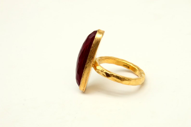 Może przedstawiać: Złoty pierścionek z dużym, fasetowanym, ciemnoczerwonym kamieniem szlachetnym. Obrączka pierścionka jest fakturowana i ma młotkowane wykończenie.