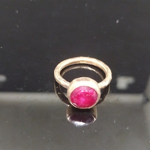 Pu&ograve; includere: Un anello d'oro con una pietra preziosa di rubino rosso. L'anello ha una texture martellata.