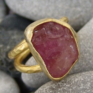 Pode incluir: Um anel de ouro com uma grande gema rosa de forma irregular. O anel tem uma banda de ouro martelada.
