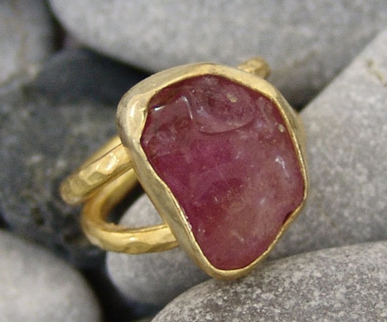 Pode incluir: Um anel de ouro com uma grande gema rosa de forma irregular. O anel tem uma banda de ouro martelada.