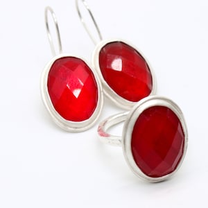 Op de afbeelding: Een set zilveren oorbellen en een ring met rode edelsteen accenten. De oorbellen zijn ovaalvormig met een zilveren haak sluiting. De ring is een zilveren band met een grote ovale rode edelsteen.