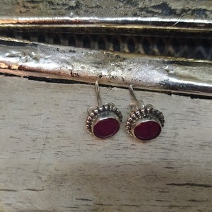 May include: A pair of silver stud earrings with small, round, dark red gemstones set in a beaded border.