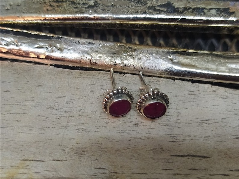 May include: A pair of silver stud earrings with small, round, dark red gemstones set in a beaded border.