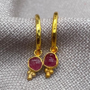 May include: A pair of gold hoop earrings featuring a hammered texture on the circular bands. Each earring suspends a single, irregular, raw red gemstone drop, accented by three small gold spheres at the base of the stone setting.