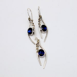 May include: A pair of silver earrings with blue gemstones and gold accents. The earrings are shaped like a teardrop with a curved line.