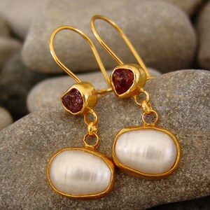 Könnte beinhalten: Goldene Ohrringe mit einem roten Edelstein und einem weißen Perlen-Tropfen. Die Ohrringe haben ein zartes Design und sind perfekt, um jedem Outfit einen Hauch von Eleganz zu verleihen.