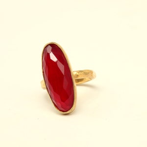 May include: A gold ring with a large, faceted, red gemstone. The ring is oval shaped and has a textured band.