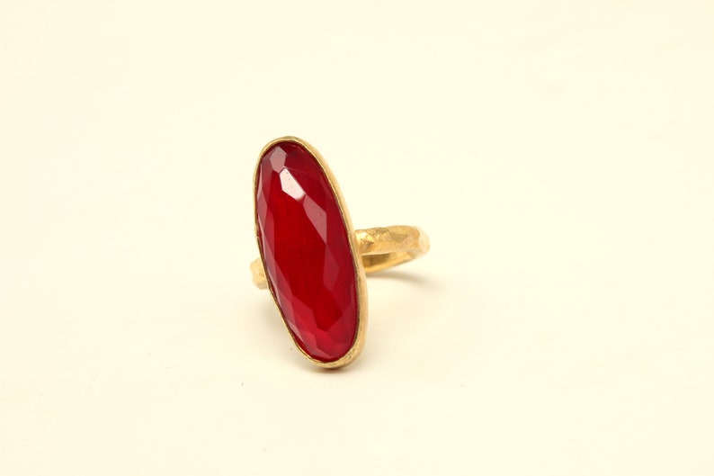 May include: A gold ring with a large, faceted, red gemstone. The ring is oval shaped and has a textured band.