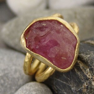 Pode incluir: Um anel de ouro com uma grande gema rosa facetada engastada em um engaste de bezel. O anel tem um design de banda tripla.