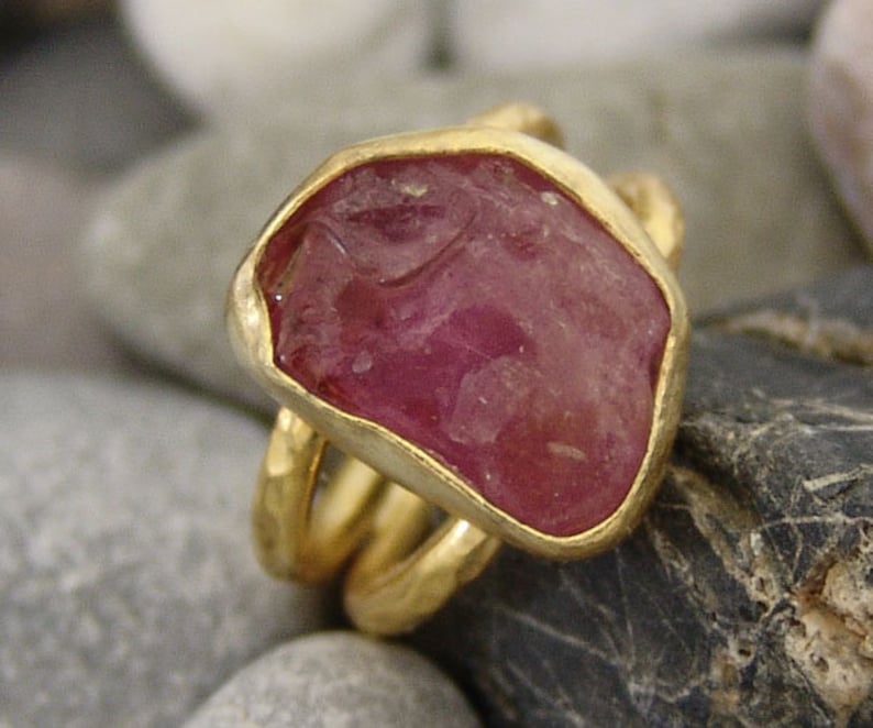 Pode incluir: Um anel de ouro com uma grande gema rosa facetada engastada em um engaste de bezel. O anel tem um design de banda tripla.