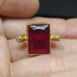 Op de afbeelding: Een gouden ring met een grote, rechthoekige, rode edelsteen en twee kleinere, ronde, witte edelstenen aan de zijkanten.