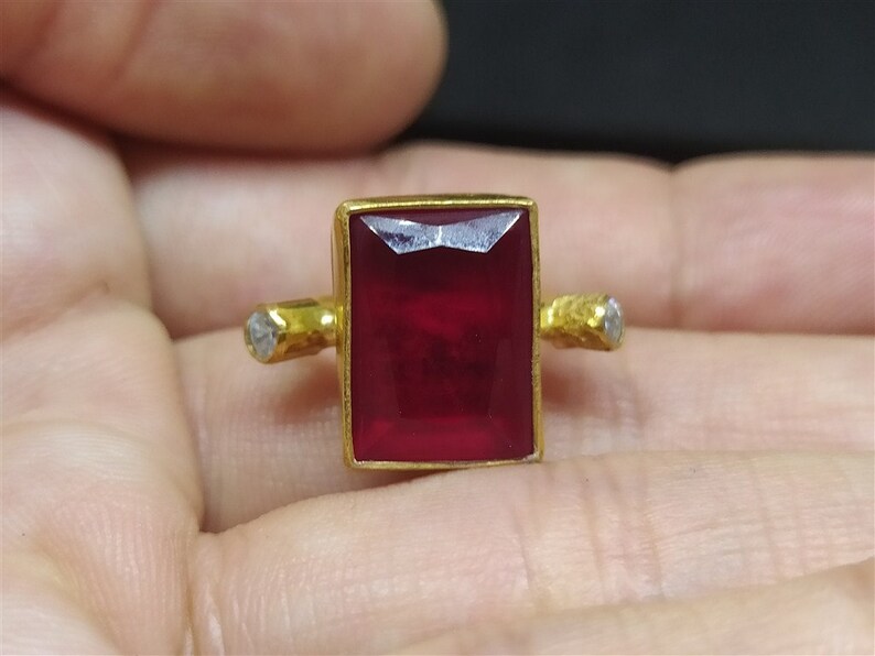 Op de afbeelding: Een gouden ring met een grote, rechthoekige, rode edelsteen en twee kleinere, ronde, witte edelstenen aan de zijkanten.