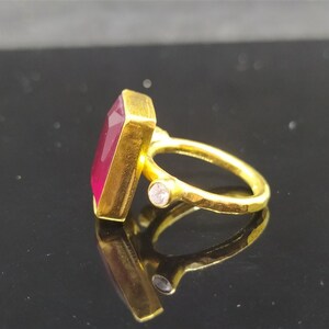 Op de afbeelding: Een gouden ring met een grote, rechthoekige, donkerrode edelsteen en een kleine, heldere edelsteen aan de zijkant.