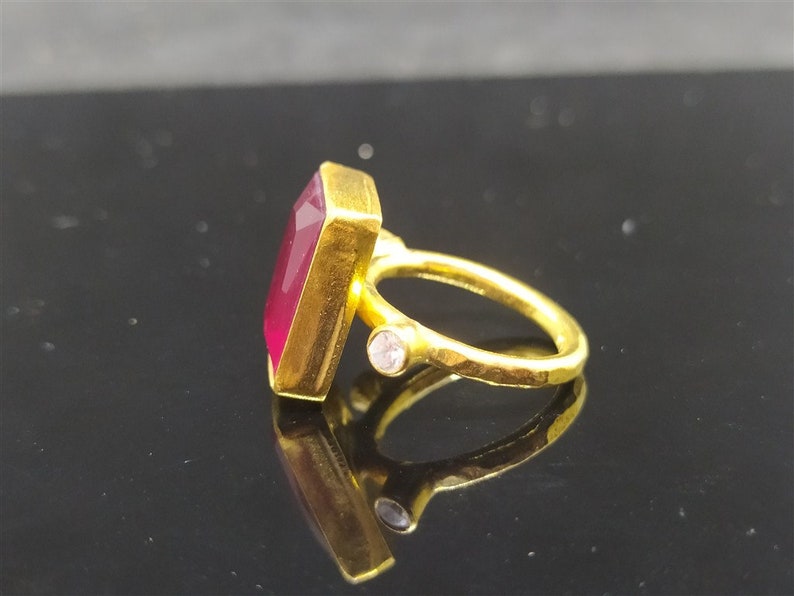 Op de afbeelding: Een gouden ring met een grote, rechthoekige, donkerrode edelsteen en een kleine, heldere edelsteen aan de zijkant.