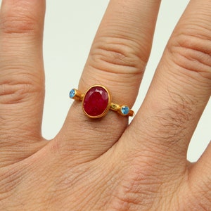 Peut inclure: Une bague en or avec une grande pierre rouge ovale et deux petites pierres bleues de chaque c&ocirc;t&eacute;.