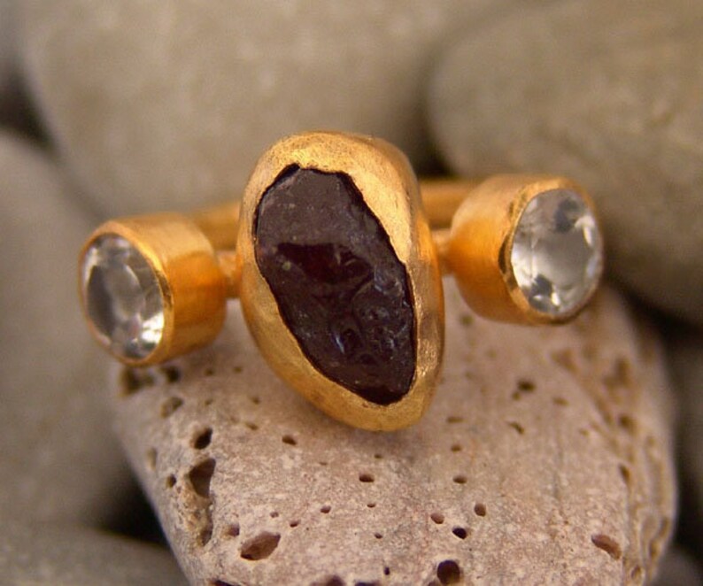 May include: Gold ring featuring a large, dark red gemstone and two smaller, clear gemstones.