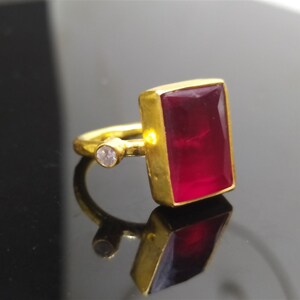 Op de afbeelding: Een gouden ring met een grote, rechthoekige, donkerrode edelsteen en een kleine, ronde, heldere edelsteen.