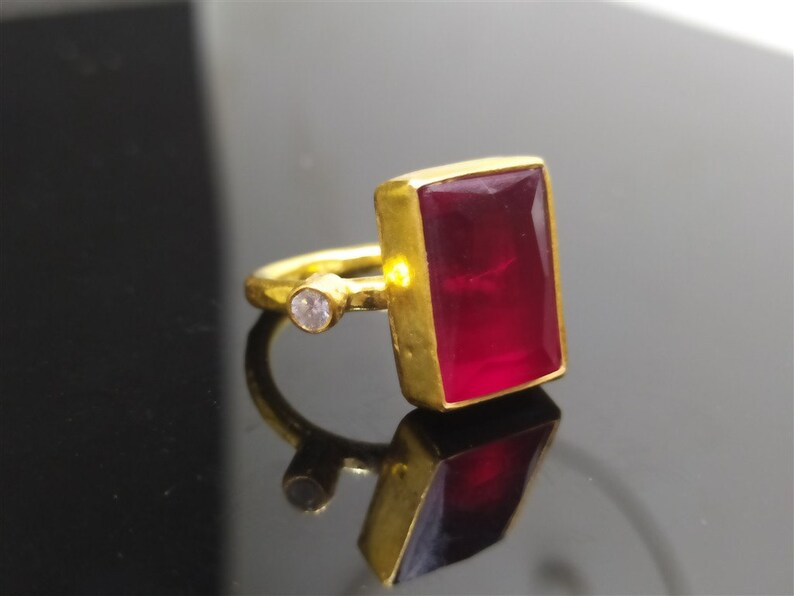 Op de afbeelding: Een gouden ring met een grote, rechthoekige, donkerrode edelsteen en een kleine, ronde, heldere edelsteen.