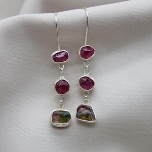 Op de afbeelding: Zilveren oorhangers met drie roze toermalijn edelstenen aan elke oorhanger. De onderste edelsteen is een grotere, veelkleurige toermalijn.