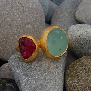 May include: A gold ring with a large, round, light blue gemstone and a smaller, heart-shaped, dark red gemstone.