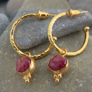 May include: Gold hoop earrings with a hammered texture and a dangling red gemstone pendant. The pendant has three small gold balls at the bottom.