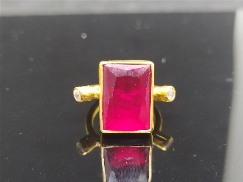 Op de afbeelding: Een gouden ring met een grote, rechthoekige, dieprode edelsteen en twee kleinere, ronde, witte edelstenen op de band.