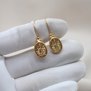 May include: A pair of gold-toned earrings with a detailed oval design. The earrings are hanging from a white surface.
