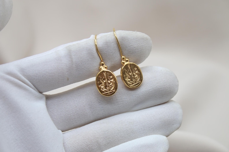 May include: A pair of gold-toned earrings with a detailed oval design. The earrings are hanging from a white surface.