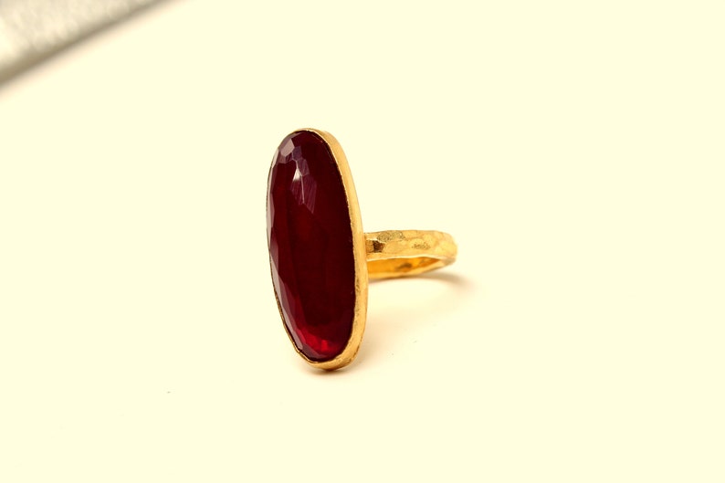 Może przedstawiać: Złoty pierścionek z dużym, owalnym, ciemnoczerwonym kamieniem szlachetnym.