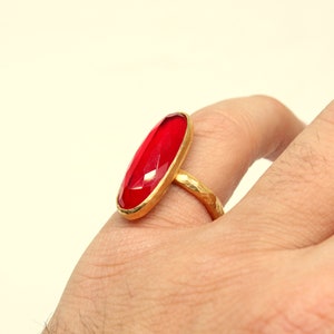 May include: A gold ring with a large, oval-shaped, red gemstone. The ring is worn on a person's finger.