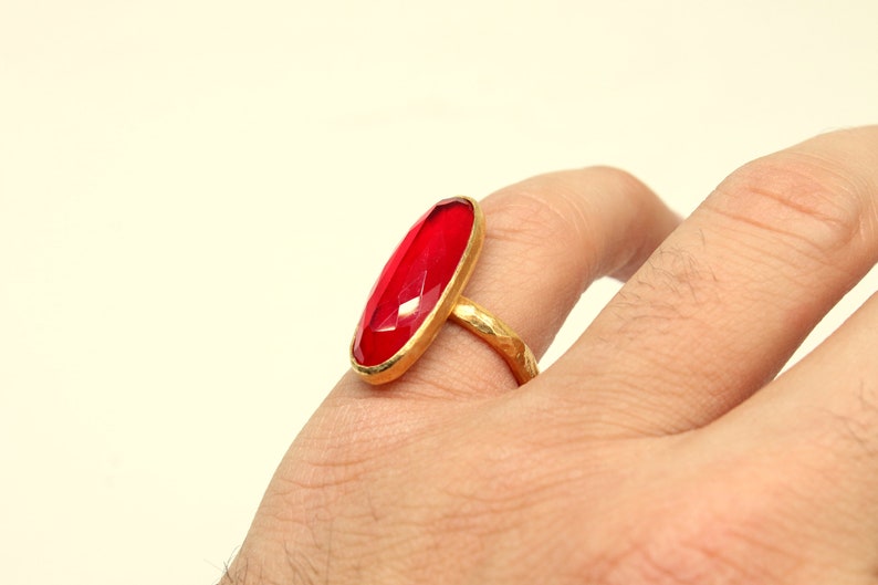 May include: A gold ring with a large, oval-shaped, red gemstone. The ring is worn on a person's finger.