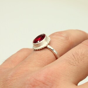 Op de afbeelding: Een zilveren ring met een rode edelsteen gezet in een getextureerde bezel. De ring wordt gedragen aan een vinger.