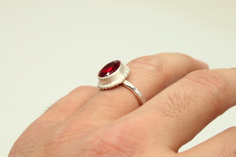 Op de afbeelding: Een zilveren ring met een rode edelsteen gezet in een getextureerde bezel. De ring wordt gedragen aan een vinger.