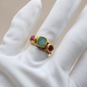 May include: A gold ring with a green gemstone and two pink gemstones. The ring is on a white surface.
