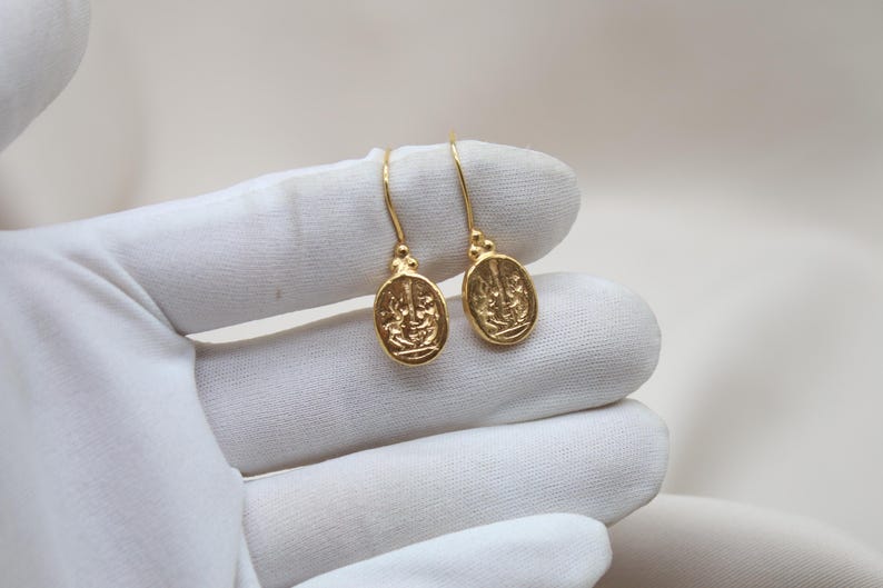 May include: A pair of gold-toned earrings with a detailed design. The earrings are oval-shaped and have a dangling design.