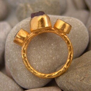 May include: A gold ring featuring three gemstones. The ring has a textured, hammered finish. The gemstones are set in gold bezels.