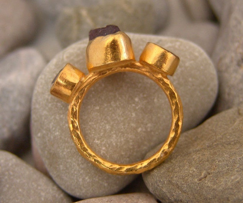 May include: A gold ring featuring three gemstones. The ring has a textured, hammered finish. The gemstones are set in gold bezels.