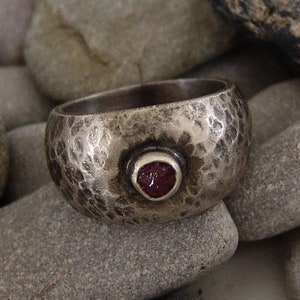 May include: A silver ring with a hammered texture and a red gemstone set in the centre.