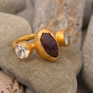May include: A gold ring featuring three stones. The central stone is a large, dark purple, irregularly shaped stone. The two smaller stones are round and clear.