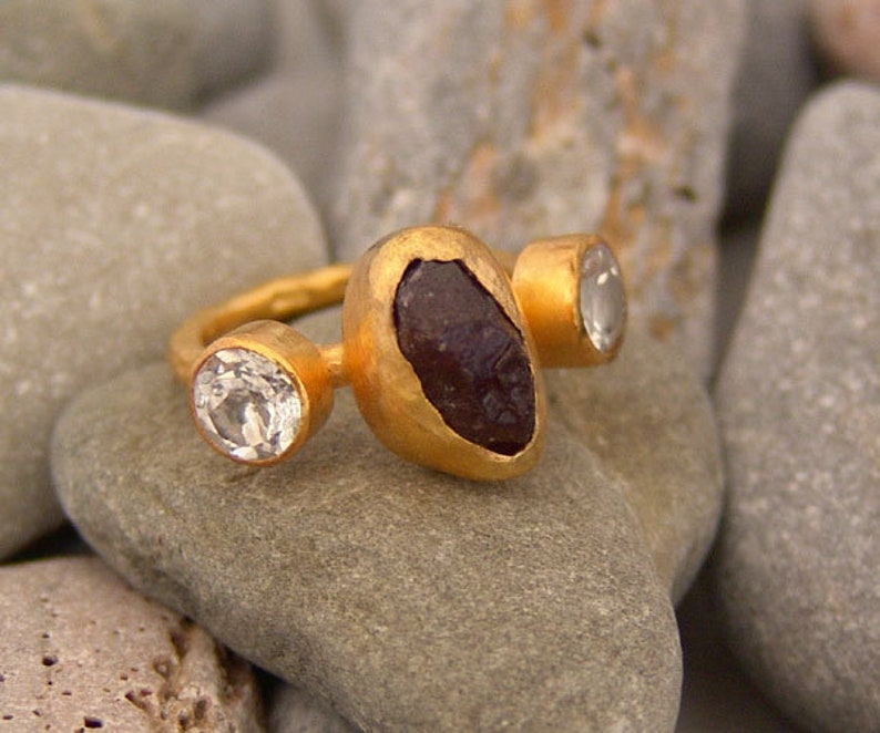 May include: A gold ring featuring three stones. The central stone is a large, dark purple, irregularly shaped stone. The two smaller stones are round and clear.