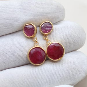 Könnte beinhalten: Ein Paar goldfarbene Ohrringe mit Rubinen. Jeder Ohrring hat zwei Steine: einen kleinen, unregelmäßigen oberen Stein und einen größeren, facettierten, kreisförmigen unteren Stein. Präsentiert auf weißem Hintergrund.