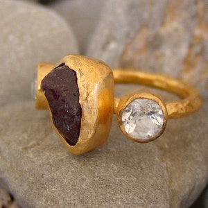 May include: A gold ring featuring a large, dark red gemstone and a smaller, clear gemstone. The ring has a hammered finish.