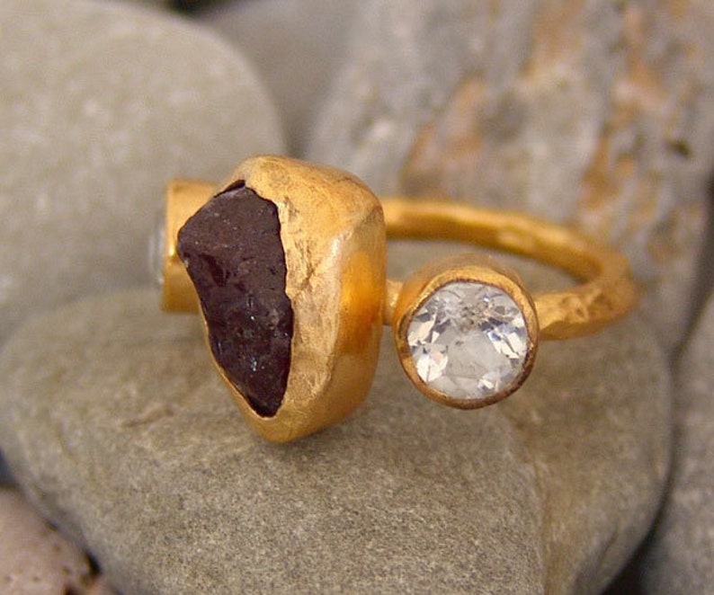 May include: A gold ring featuring a large, dark red gemstone and a smaller, clear gemstone. The ring has a hammered finish.