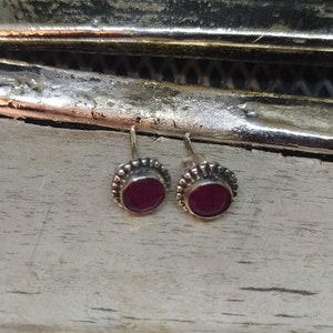 May include: A pair of silver stud earrings with small, round, dark red gemstones set in a beaded border.