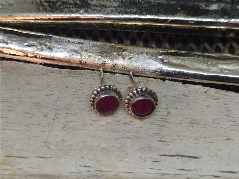 May include: A pair of silver stud earrings with small, round, dark red gemstones set in a beaded border.