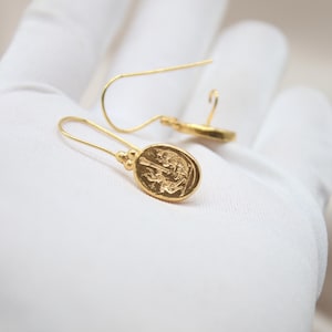 May include: A pair of gold dangle earrings with a detailed oval pendant featuring a raised design. The earrings have a simple, elegant style.