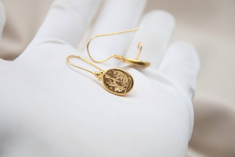 May include: A pair of gold dangle earrings with a detailed oval pendant featuring a raised design. The earrings have a simple, elegant style.
