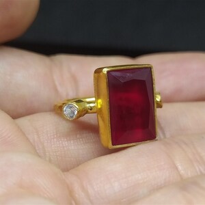 Op de afbeelding: Een gouden ring met een grote, rechthoekige, rode edelsteen en een kleinere, ronde, heldere edelsteen.