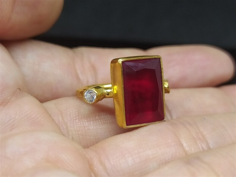 Op de afbeelding: Een gouden ring met een grote, rechthoekige, rode edelsteen en een kleinere, ronde, heldere edelsteen.