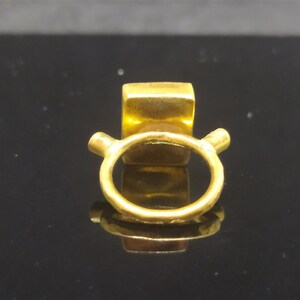 Op de afbeelding: Een gouden ring met een vierkante steen in het midden. De ring heeft een eenvoudige band en twee kleine steunen aan weerszijden van de steen.